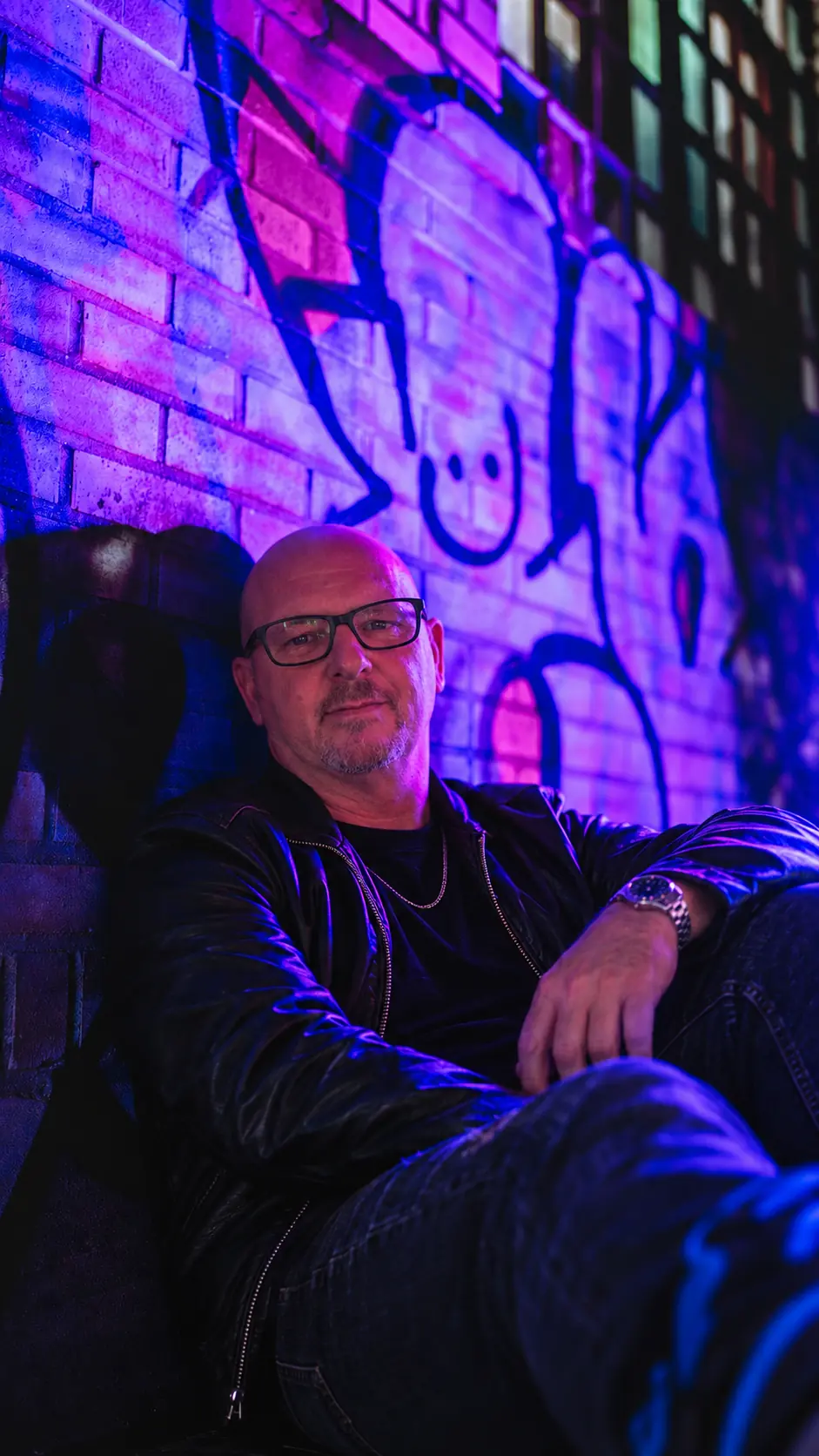 DJ Bill Bass seated against a neon-lit graffiti wall in Downtown Austin